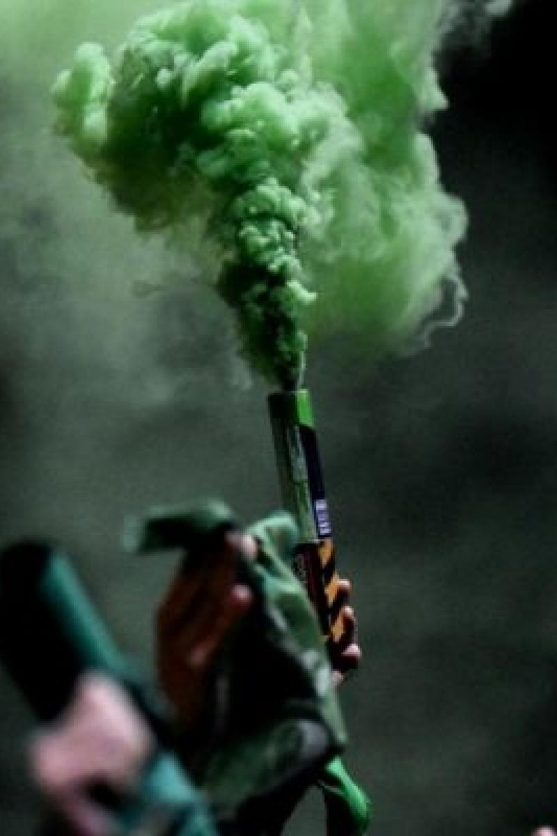 Esta es una imagen. En la imagen se muestra una mano sosteniendo un cilindro del que sale humo color verde.